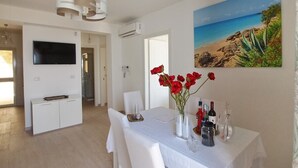 A modern dining area with a white table, chairs, and a vase of red flowers. There is a painting of a beach on the wall and a flat-screen TV mounted above the table.