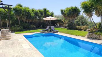 A rectangular swimming pool with a clear blue water surface, surrounded by a stone deck. There are two lounge chairs with umbrellas, and a small staircase leading to a covered area.