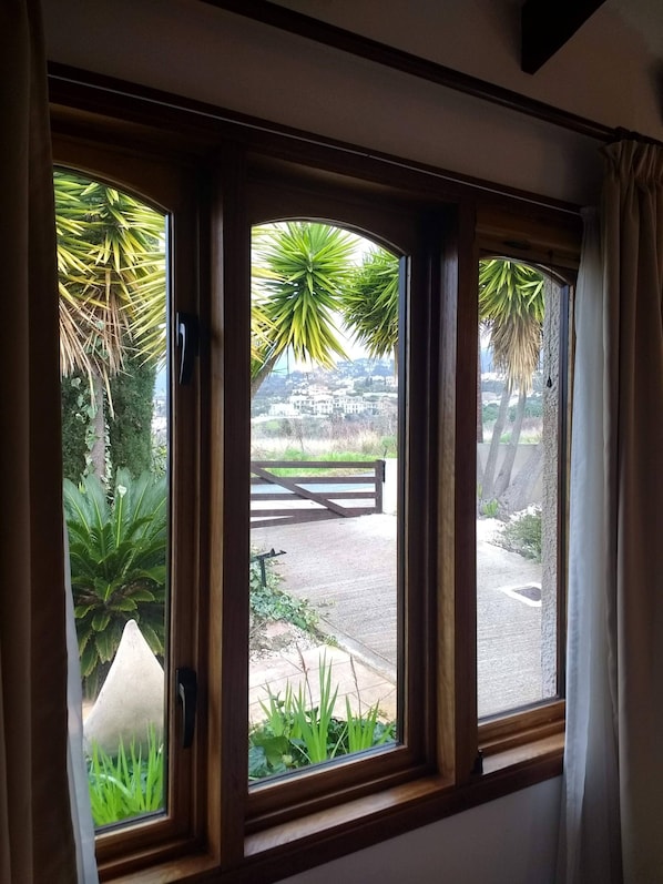 A room with three windows overlooking a garden and a distant view of buildings. - A charming villa set in beautiful gardens with sea & mountain views  (Paphos)