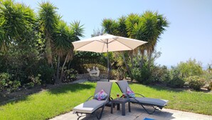 Zona junto a la piscina con dos sillones de descanso y una mesa bajo una pérgola, rodeada de abundante vegetación y una piscina azul cristalina.
