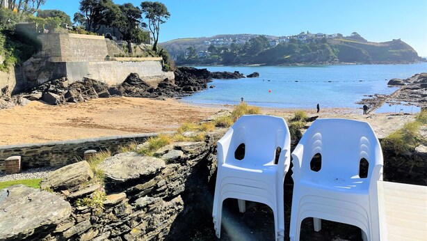 A beach with rocky terrain, a stone wall, and white plastic chairs. - Seaweed Cottage in Fowey overlooking the beach with a garden and a parking space (Fowey)