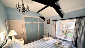 A bedroom with a large bed, a bedside table, a lamp, a window with curtains, and a chandelier. - Seaweed Cottage in Fowey overlooking the beach with a garden and a parking space (Fowey)