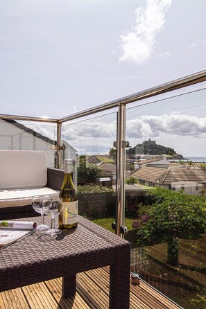 Ein Balkon mit einem Korbstuhl-Tisch, einer Weinflasche und Gläsern. Durch das Glasgeländer ist eine malerische Aussicht auf eine Küstenstadt und eine Burg zu sehen.