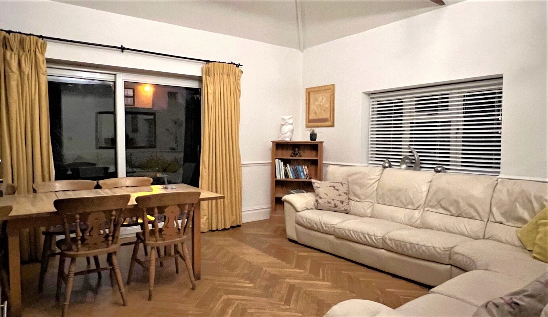 Een woonkamer met een bank, een eettafel met stoelen, een boekenkast en een raam met zonwering.