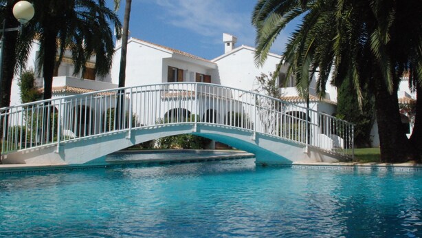 A bridge over a swimming pool with a white building and palm trees in the background.