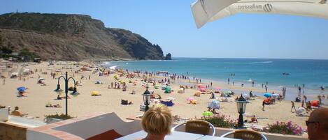 Un café frente a la playa con terraza al aire libre, con vista a una playa concurrida y al océano.