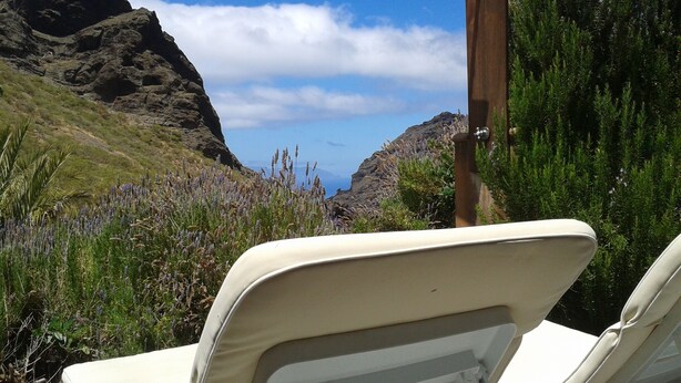 A beachside view with a mountain in the background, a wooden post with a solar panel, and two beige lounge chairs. - Enjoy Casa Rural La Tinta (Buenavista del Norte)
