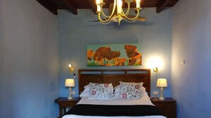 A bedroom with a wooden headboard, a bed with white bedding, two bedside lamps, a painting of flowers on the wall, and a chandelier. - Enjoy Casa Rural La Tinta (Buenavista del Norte)