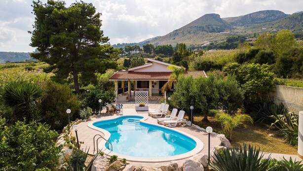 A pool area with lounge chairs and a house in the background.