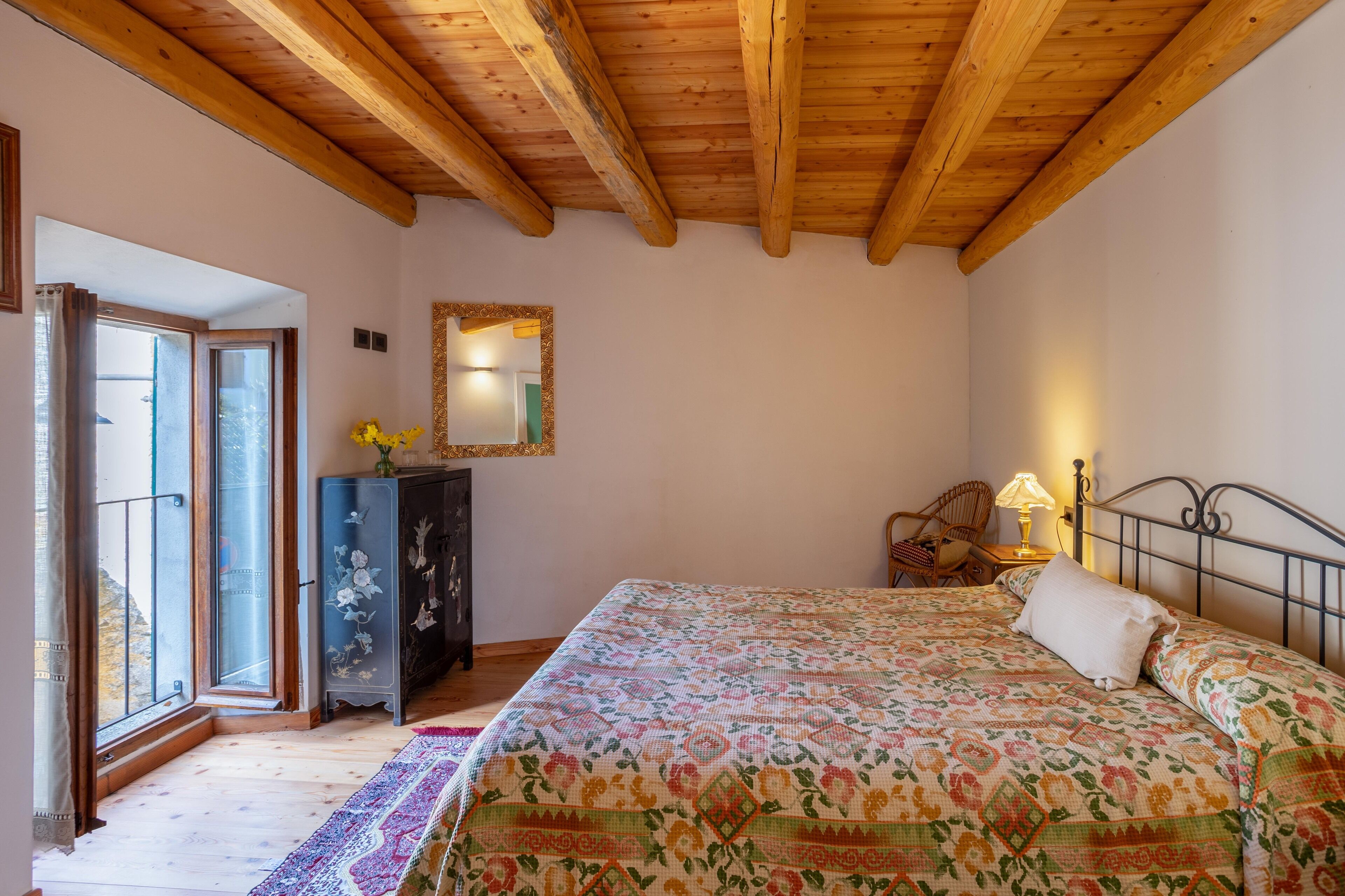 Une chambre avec un plafond en bois, un lit recouvert d’une couvre-lit à motifs floraux, une commode, un miroir et une fenêtre avec des rideaux.