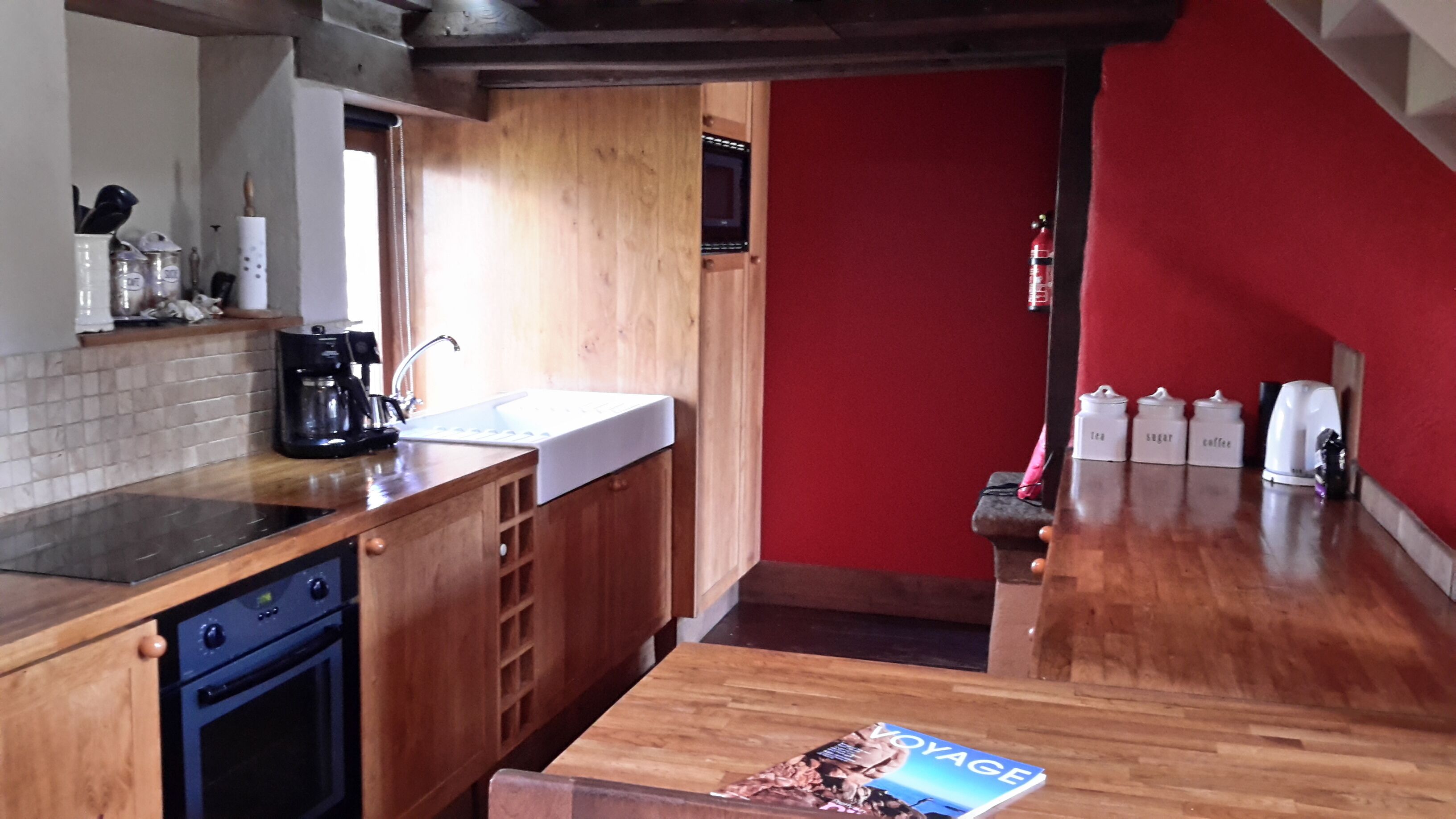 Una cocina con armarios de madera, un lavavajillas negro, un lavabo blanco y una pared roja. Hay cafeteras y una revista en la encimera.