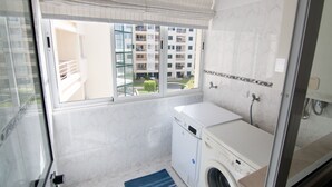 Een kleine hotelbadkamer met een wasmachine, een douche en een raam met uitzicht op een woongebouw.