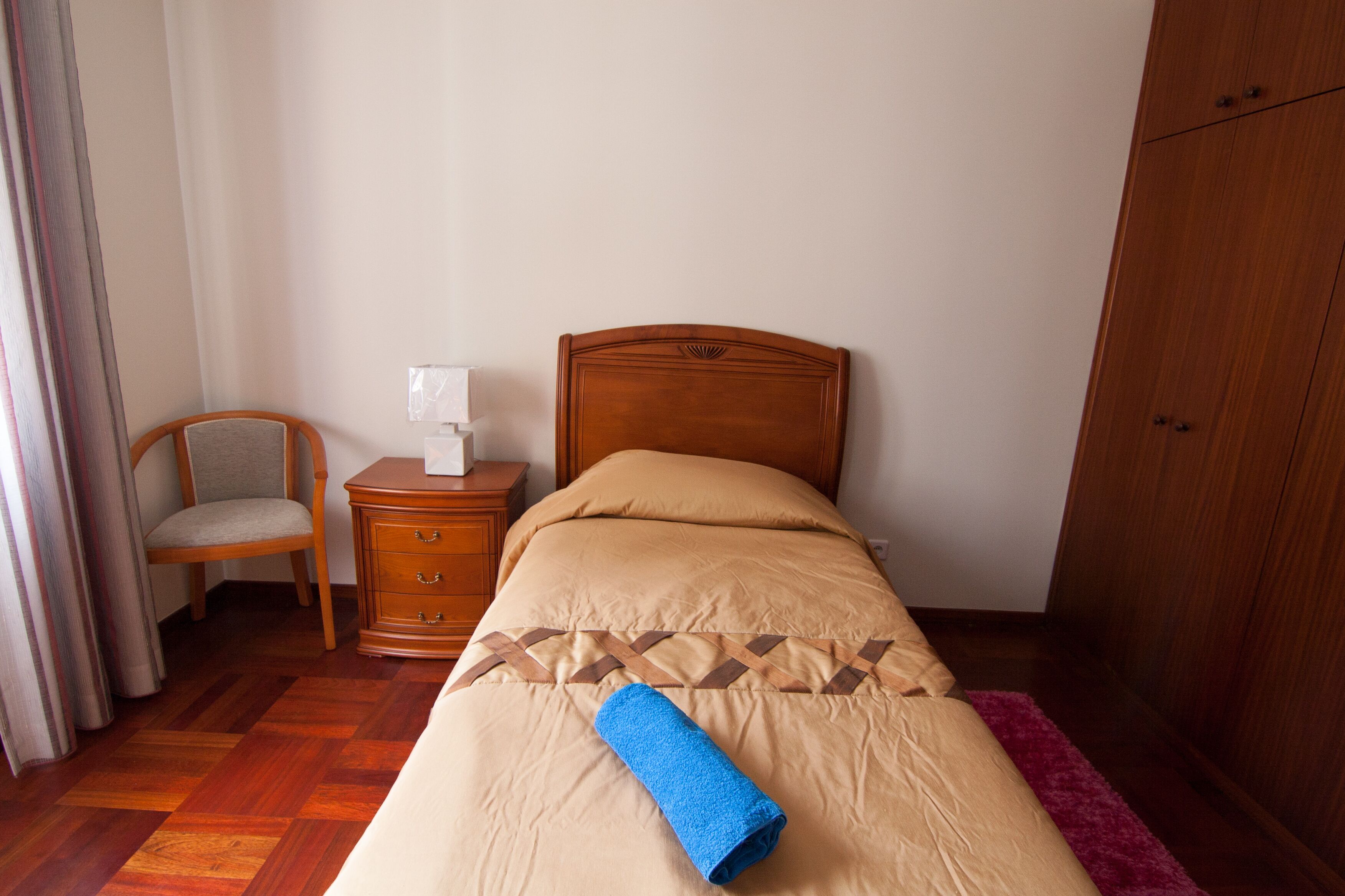 Ein ordentlich bezogenes Bett mit brauner Kopfteil, ein Nachttisch aus Holz mit Lampe, ein Stuhl und ein Kleiderschrank.