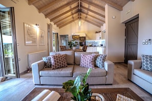 A living room with a high wooden ceiling, a sofa set, a coffee table, and a kitchen in the background.
