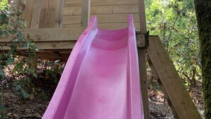 Une cabane en bois avec un toboggan rose, entourée d’arbres et de verdure.