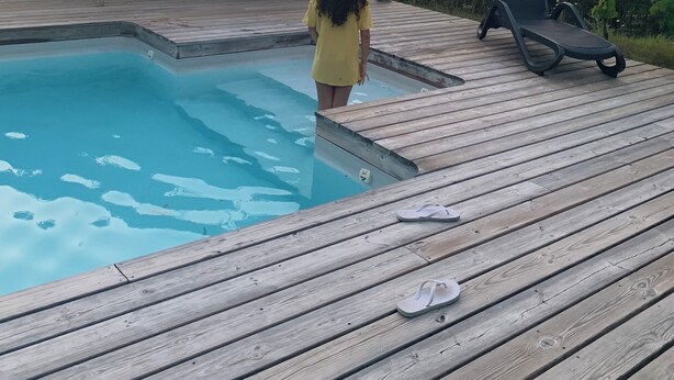 Une terrasse en bois avec une piscine, une chaise longue et un enfant debout près de la piscine.