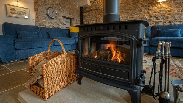A cozy living room with a stone wall, a blue sofa, a wicker basket, and a wood-burning stove.