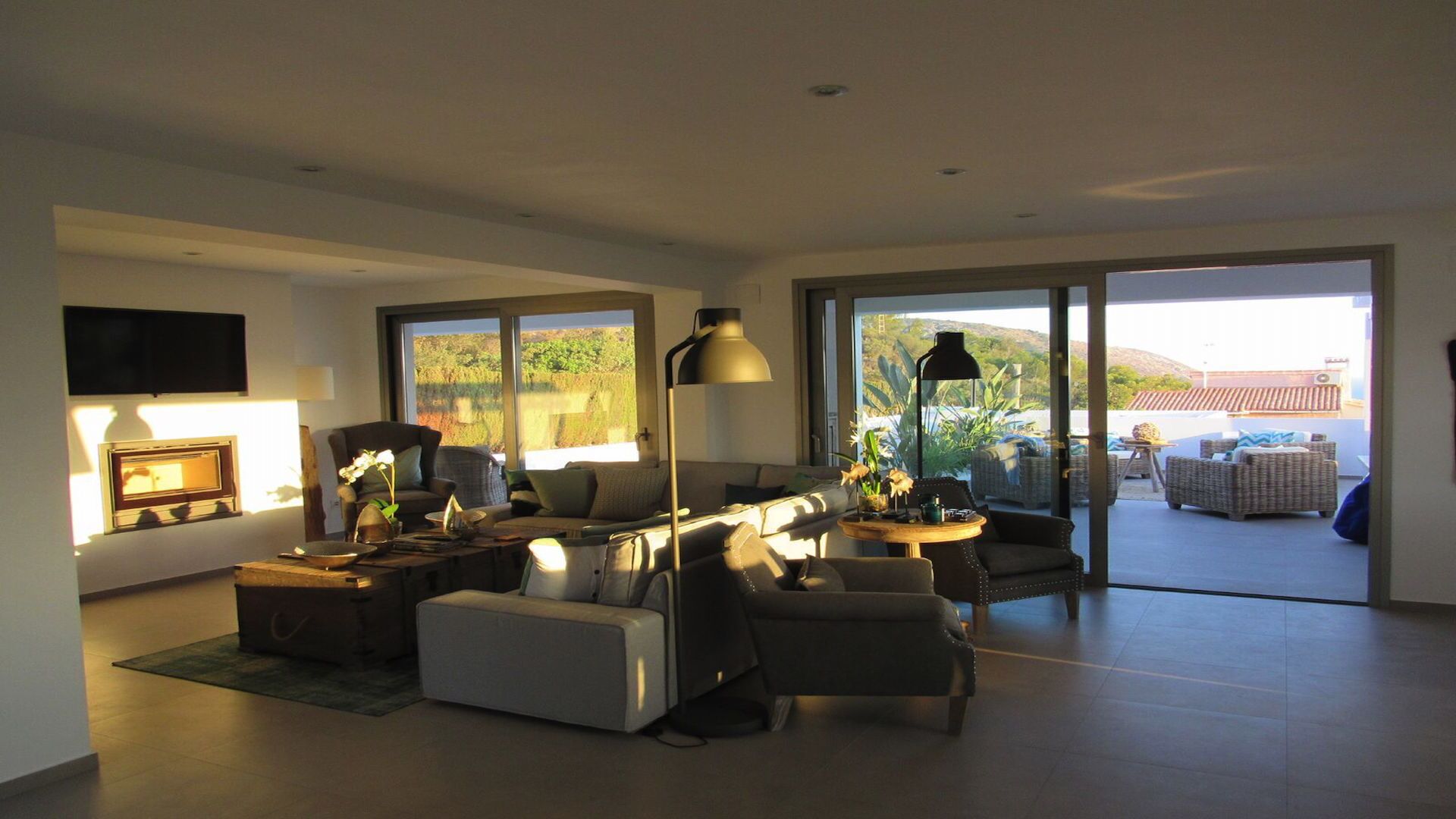 Un salon moderne avec un canapé, des fauteuils et une table basse. Un téléviseur est fixé au mur, il y a une grande fenêtre et une vue sur l’extérieur.