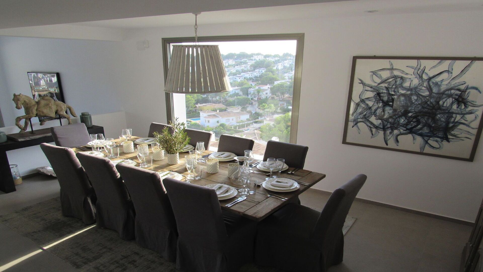 Un coin repas avec une table dressée pour six personnes, une grande fenêtre donnant sur un quartier résidentiel et un tableau abstrait moderne accroché au mur.