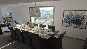 A dining area with a table set for six, a large window with a view of a residential area, and a modern abstract painting on the wall. - Villa Veintidos Large Luxury Villa Sleeps 12 (El Portet, Moraira)