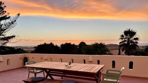 An outdoor patio with a wooden table and chairs, a bench, and a view of the landscape during sunset. - Quinta da Liberdade: studio 3 (with pool view) (Montinhos Da Luz)