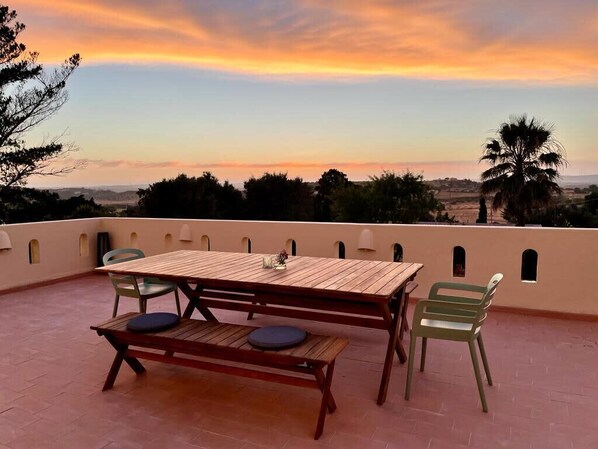 An outdoor patio with a wooden table and chairs, a bench, and a view of the landscape during sunset.