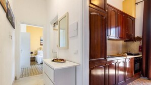 Un estrecho pasillo que da a un dormitorio con una cama y una mesita pequeña. La habitación tiene piso de baldosas y una pared blanca.
