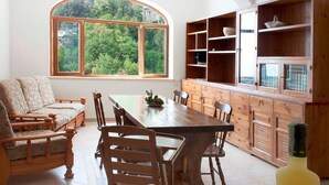 Zona pranzo con tavolo e sedie in legno, un divano e un'ampia finestra vista alberi. Sul tavolo c'è una ciotola di frutta e una bottiglia di vino.
