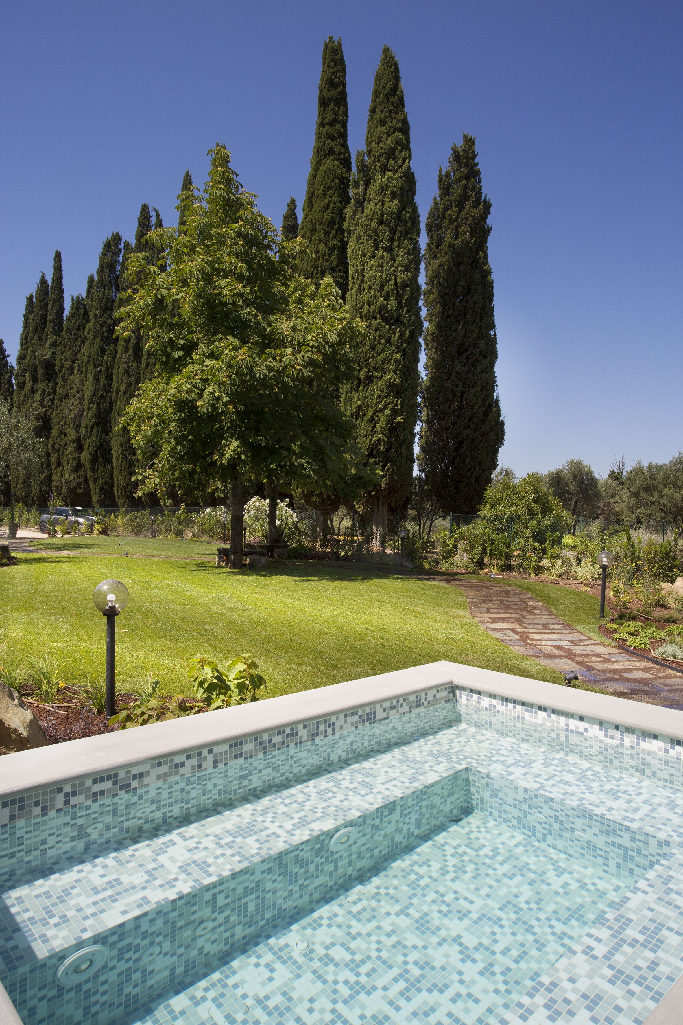 Une piscine dont le bord est recouvert de carreaux, entourée d’une pelouse bien entretenue et de grands arbres.