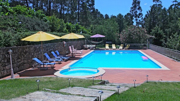 Una piscina rectangular con un muro de piedra y un borde empedrado. Se observan sillones de descanso y sombrillas alrededor de la piscina.
