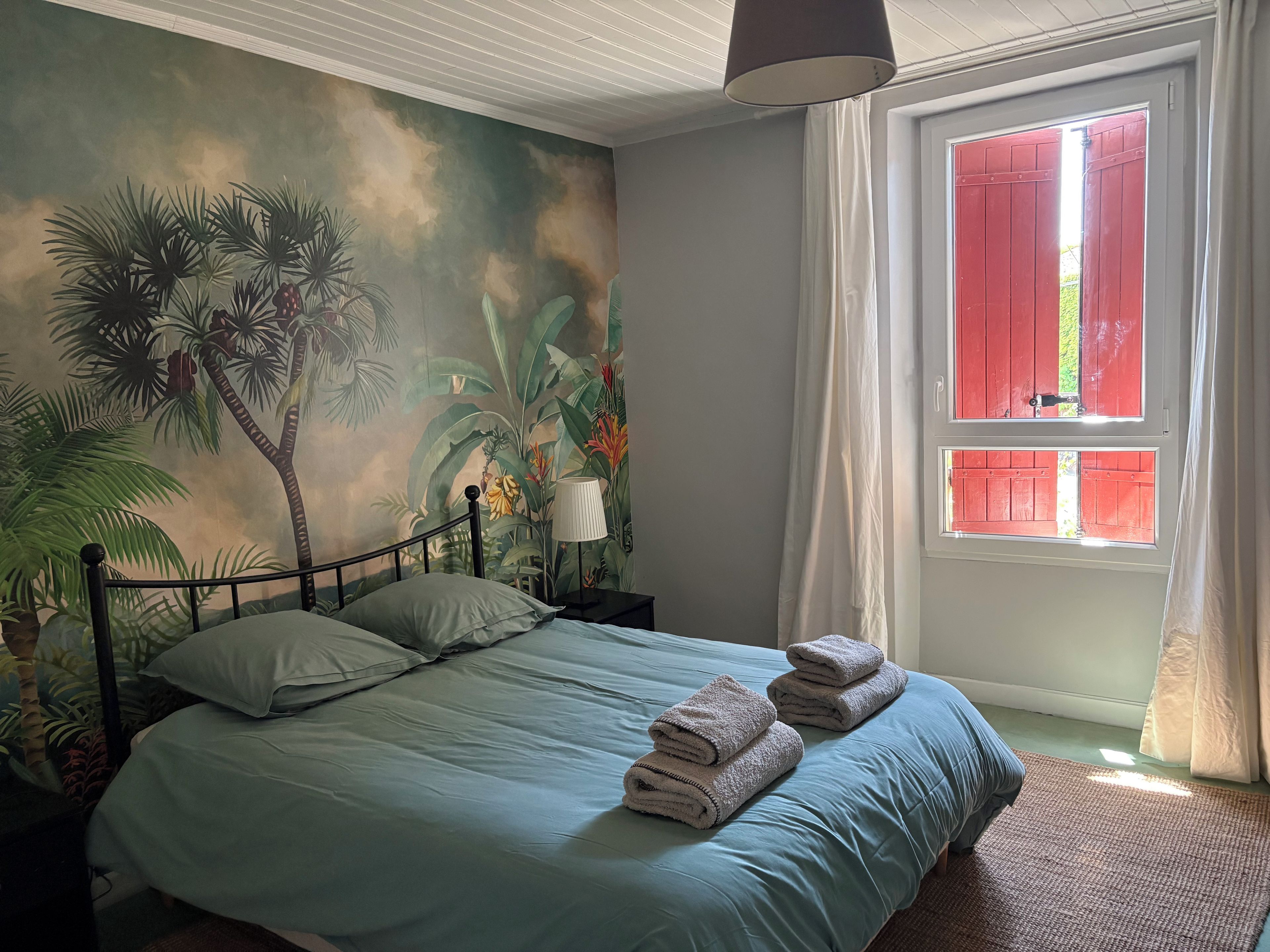 A bedroom with a bed, a nightstand, a lamp, a window with red shutters, and a tropical mural on the wall.