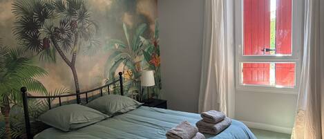 A bedroom with a bed, a nightstand, a lamp, a window with red shutters, and a tropical mural on the wall.