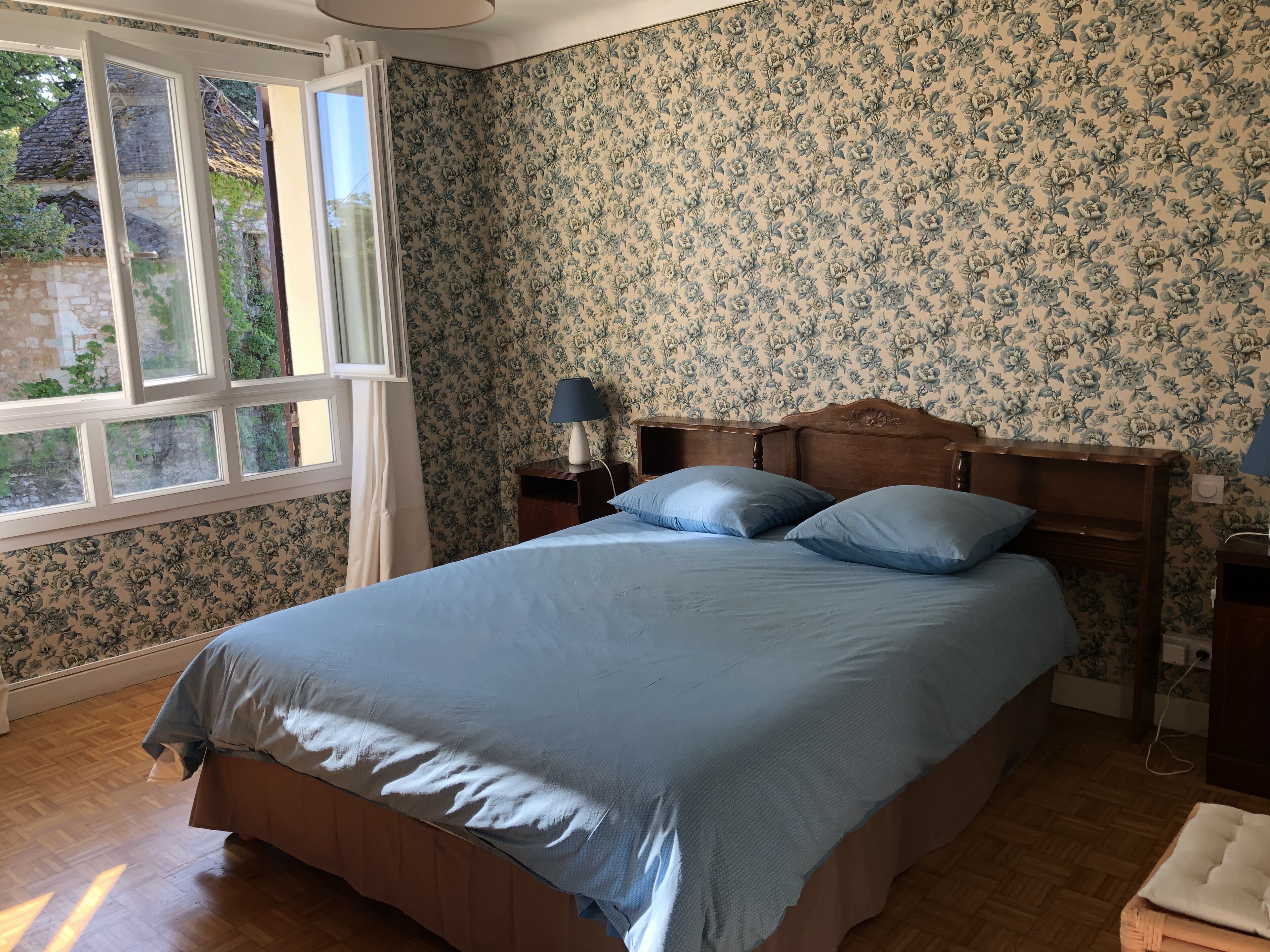 Un dormitorio con una cama de madera, papel tapiz floral, una mesita de noche con lámpara y una ventana con cortinas.
