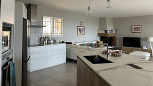 Una cocina moderna con una isla central, armarios blancos y un horno empotrado. Hay una zona de comedor con un sofá blanco y una chimenea.