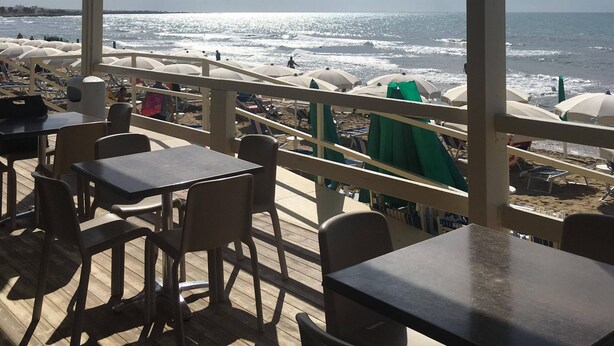 Een overdekt terras met tafels en stoelen, met uitzicht op een strand met parasols en mensen.
