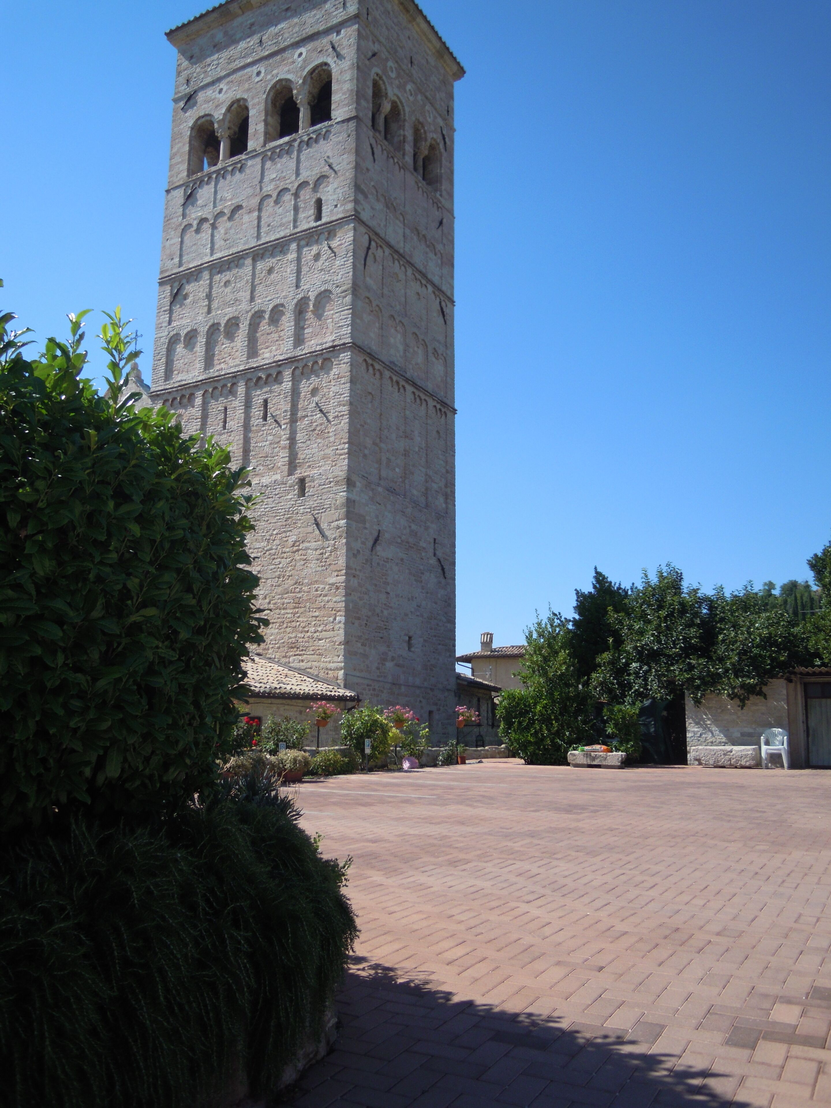 A historic stone tower with multiple arched windows, surrounded by a paved courtyard with potted plants and a clear blue sky.
