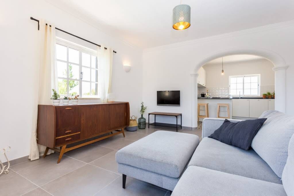 Una sala moderna con un sofá, un mueble de madera, un televisor y una cocina visible a través de un arco.