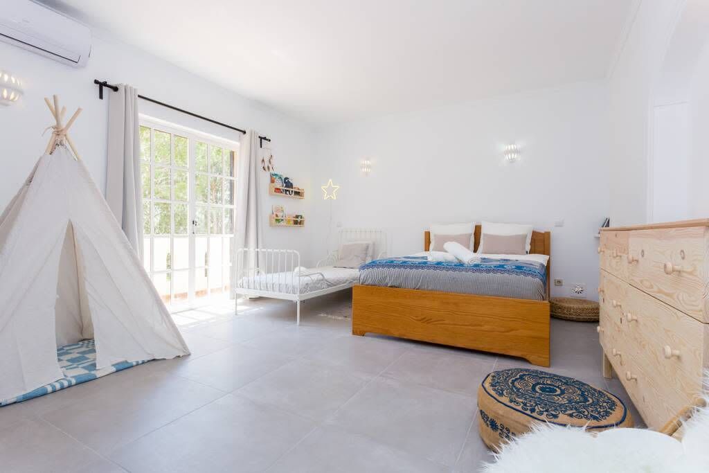 A bedroom with a bed, a white chair, a wooden dresser, a white wardrobe, and a tent.