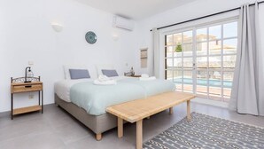 A bedroom with a bed, bench, bedside table, and a view of a swimming pool.