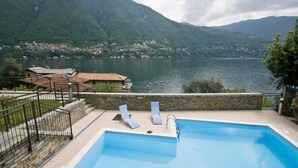 A swimming pool with lounge chairs and a stone wall, overlooking a lake and mountains.