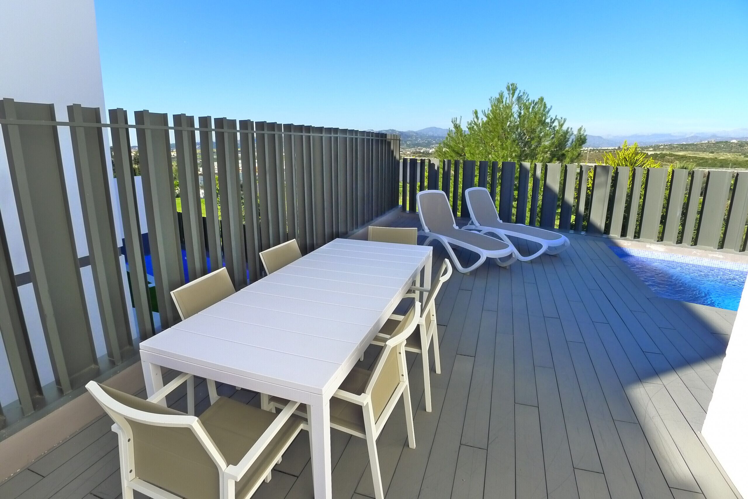 Un patio al aire libre con una mesa y sillas blancas, una piscina y vistas a montañas distantes.
