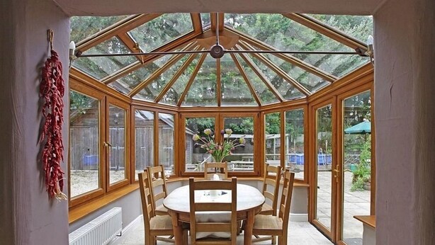 A conservatory with a wooden table and chairs, large windows, and a view of the outdoors.