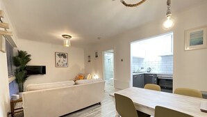 A modern living room with a sofa, dining table, and chairs. There is a kitchen area visible through an open door. - Westward 14, Polzeath, North Cornwall  (Polzeath)