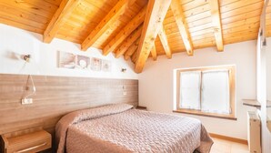A bedroom with a wooden ceiling, a bed with a quilted cover, a bedside table, a wall-mounted light, and a window with curtains.