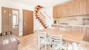 A modern kitchen with wooden cabinets and a central island, a dining area with a wooden table and chairs, and a staircase leading to an upper level.
