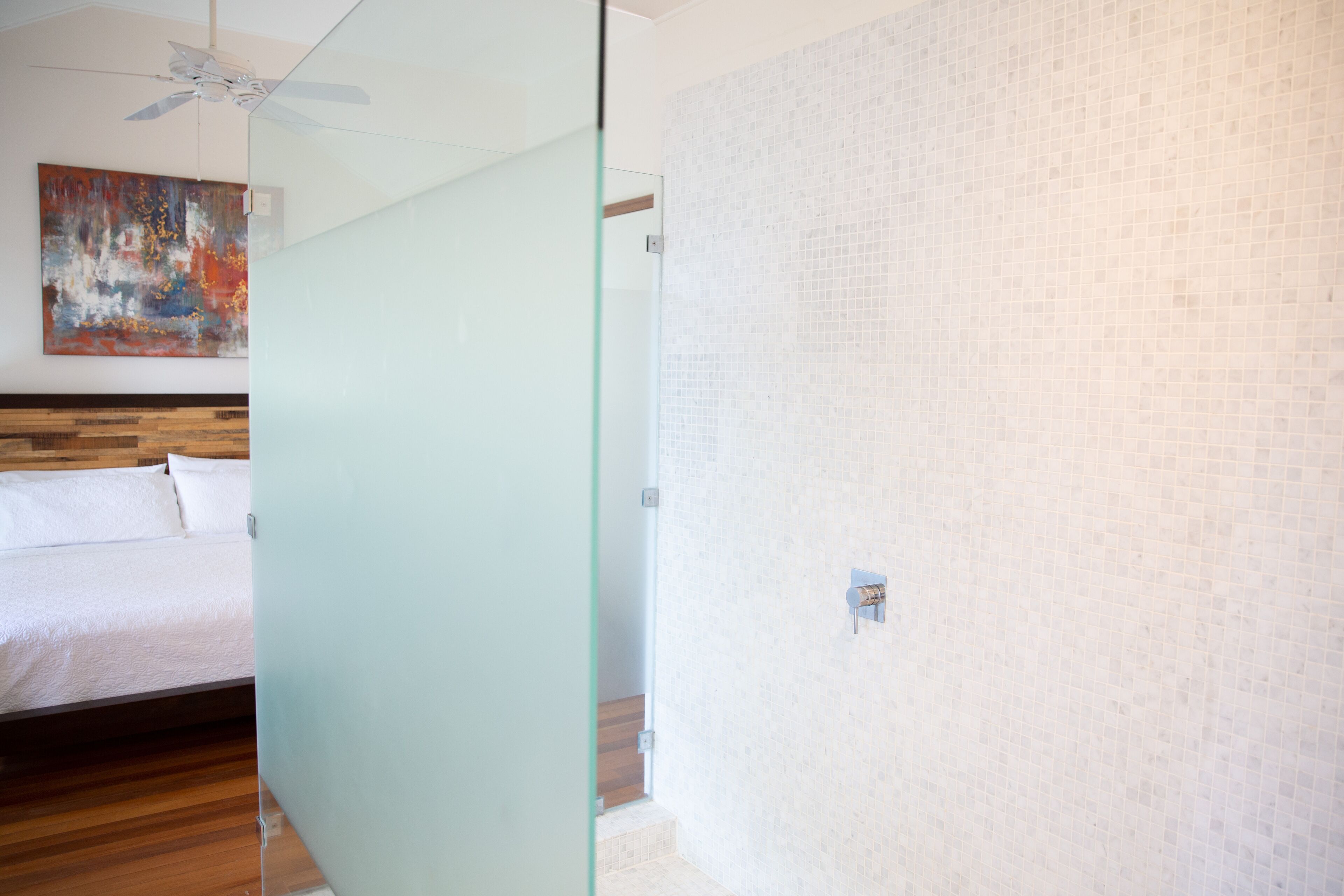 Una habitación de hotel moderna con suelo de madera, una cama con sábanas blancas, un separador de vidrio, un ventilador de techo y una pared con azulejos.