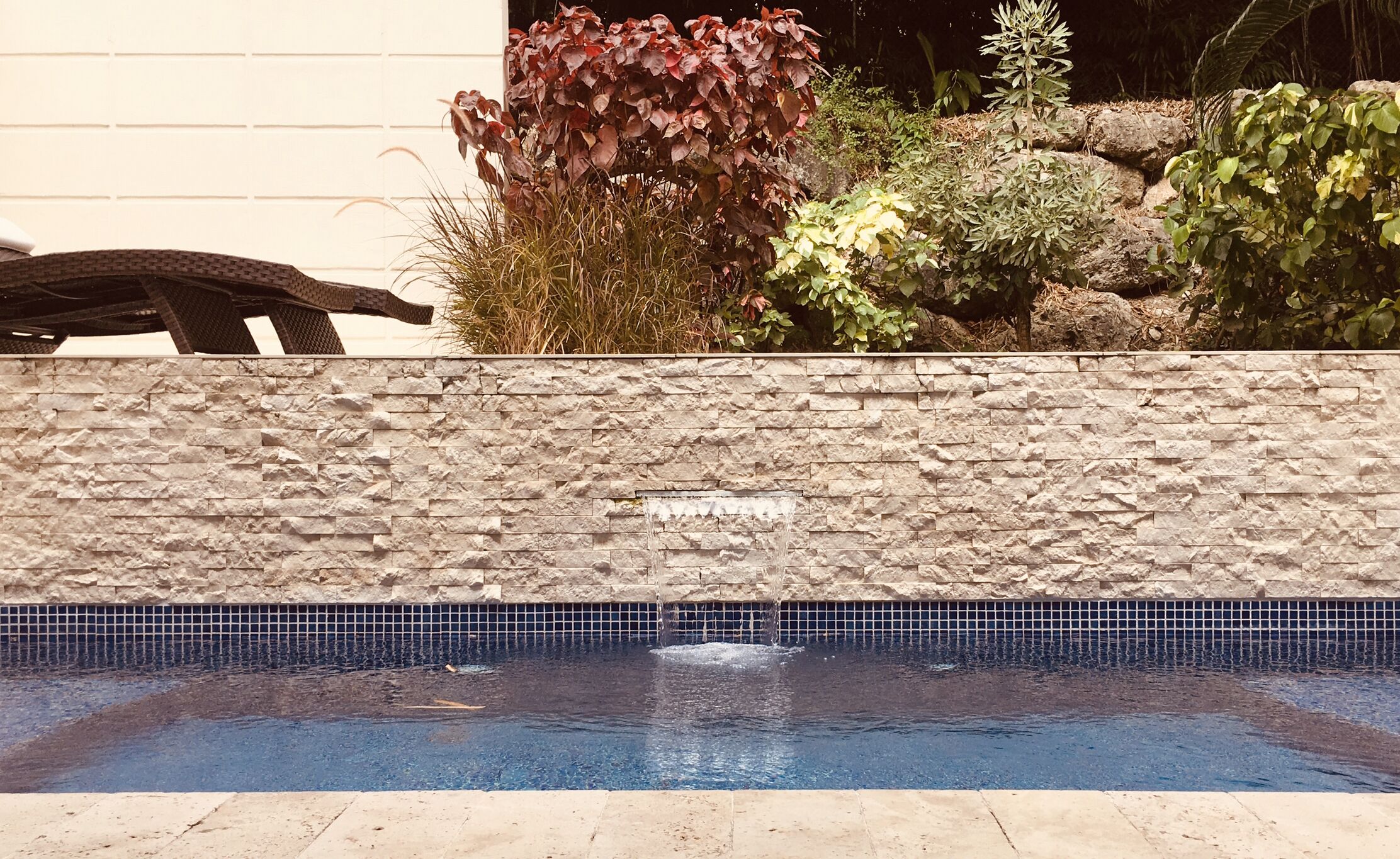 En swimmingpool med en mur av sten och en kant av kakel, omgiven av olika växter och en bänk.