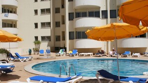 Zona de piscina con sillas reclinables azules y blancas, sombrillas amarillas y un edificio con varios balcones al fondo.