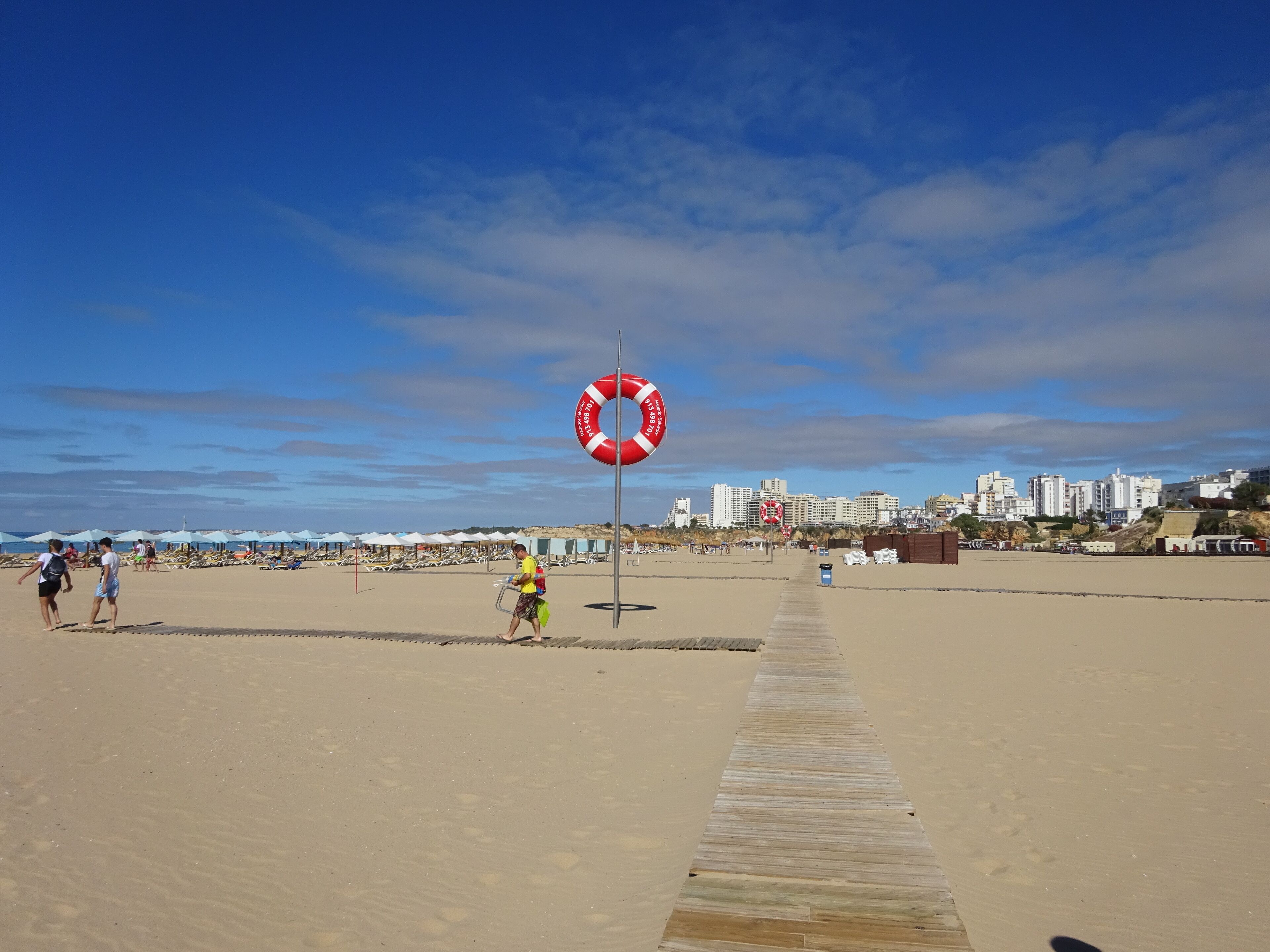Una platja amb un bóia salvavides, una passera de fusta i un paisatge urbà al fons.