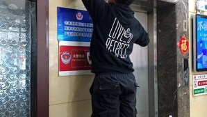 A person standing in a hallway, wearing a black hoodie with "LIVING PERFECT" on the back. There are signs on the wall, one of which is red with white text and a symbol, and another is blue with white text. - Nongjun Hotel (Beijing)